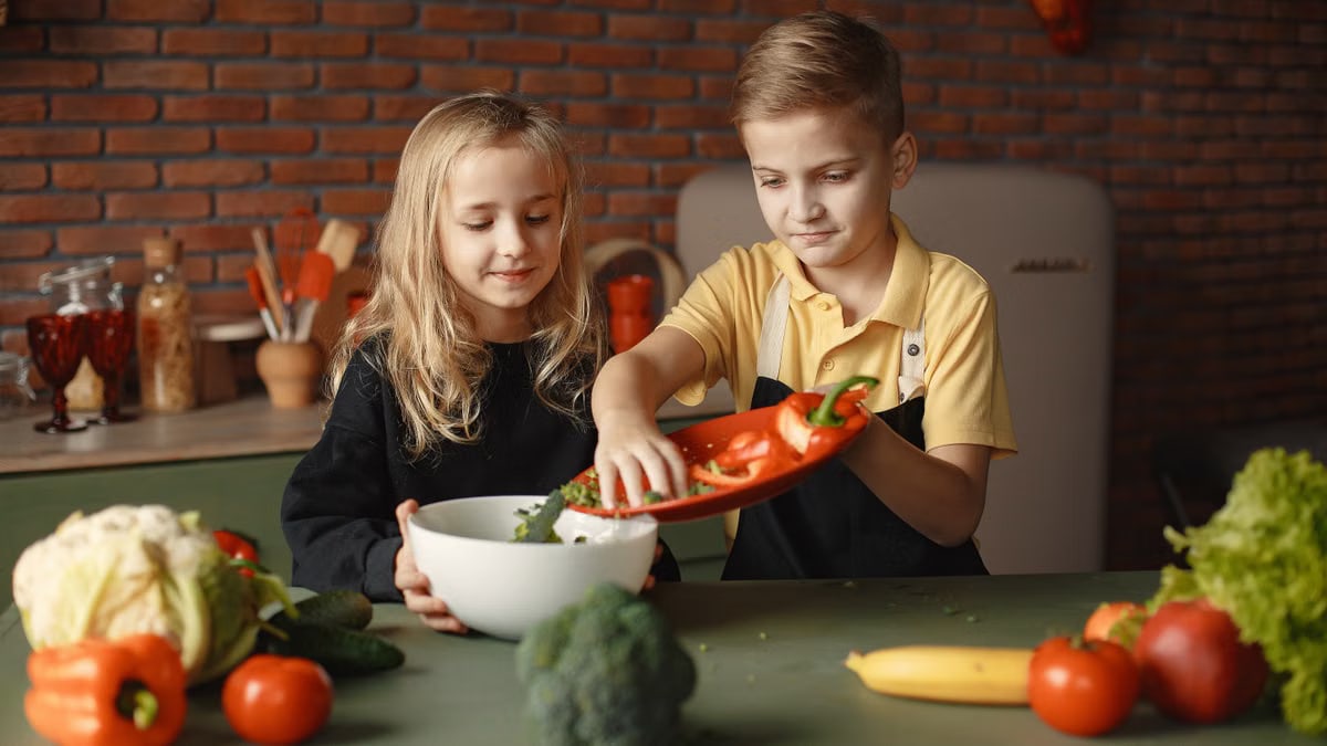 Little girl and boy mixing cutted pepper with other fresh ingredients for salad in big bowl while cooking at table with various fresh vegetables and fruits